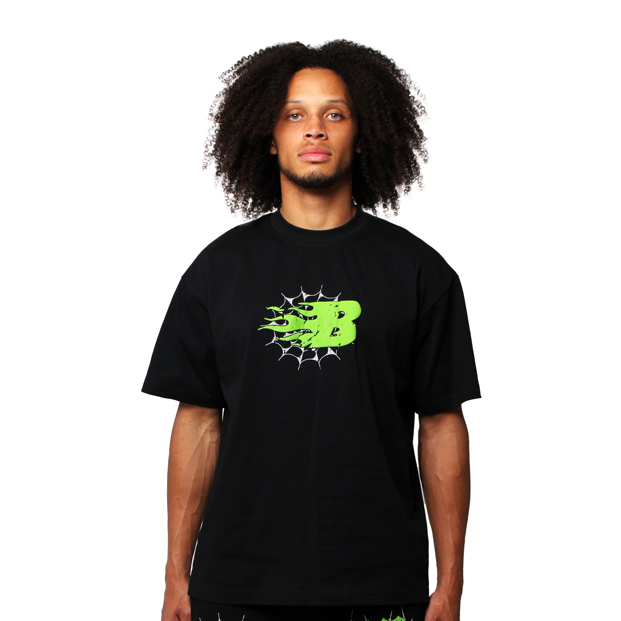 A person with curly hair wears the BEAT THE ODDS S/S TEE BLACK, featuring a neon green cosmic graphic and white web-like lines on the chest, standing facing forward against a plain white background.