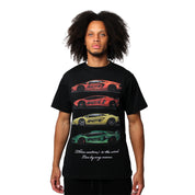 A person with medium-length curly hair wears the CAUTION TO THE WIND S/S TEE BLACK, an oversized heavy GSM cotton tee with puff-printed orange, yellow, and green sports cars stacked vertically and white cursive text below. White background.
