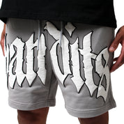 A person wears EGO SWEATSHORT GREY, featuring heavyweight French Terry Fleece and bold white and black Old English-style 3D puff print lettering across the front. Their hands are at their sides, with a black shirt partially visible.