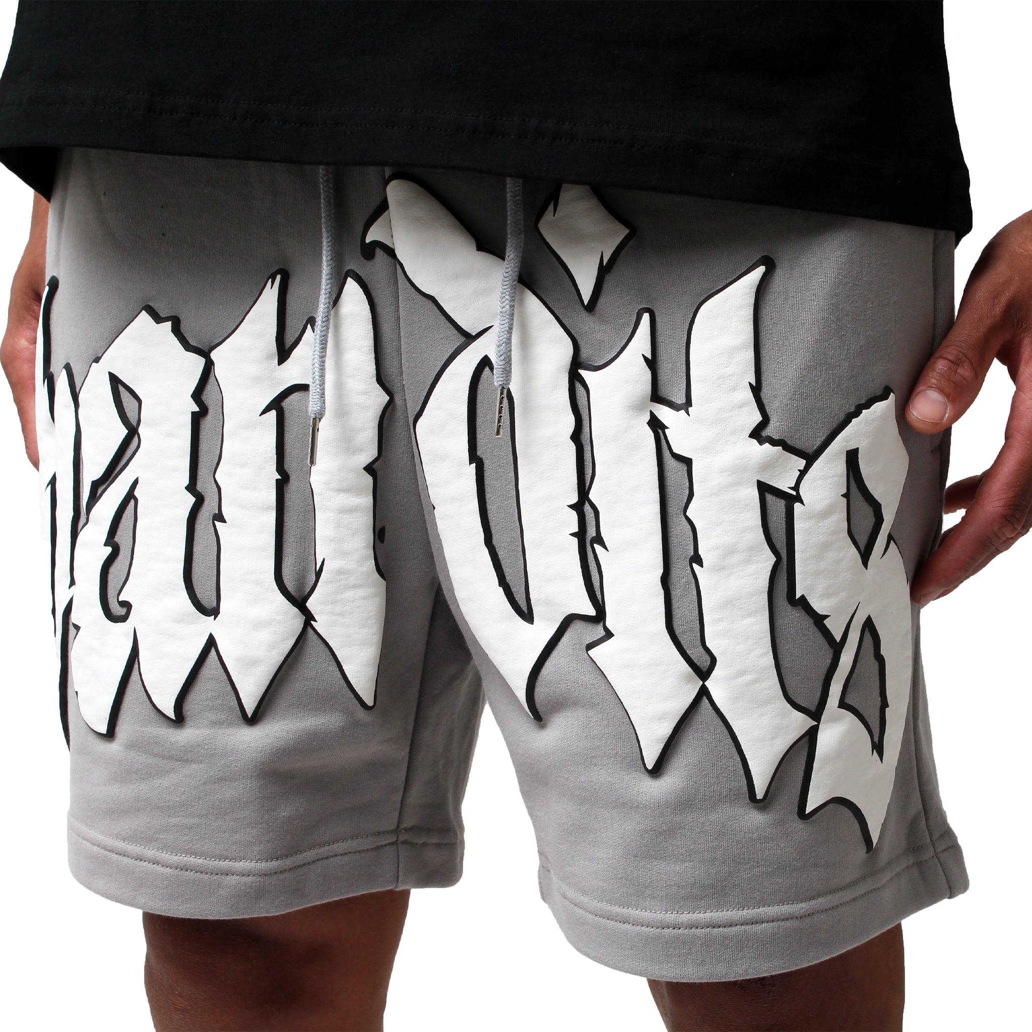 A person wears EGO SWEATSHORT GREY, featuring heavyweight French Terry Fleece and bold white and black Old English-style 3D puff print lettering across the front. Their hands are at their sides, with a black shirt partially visible.