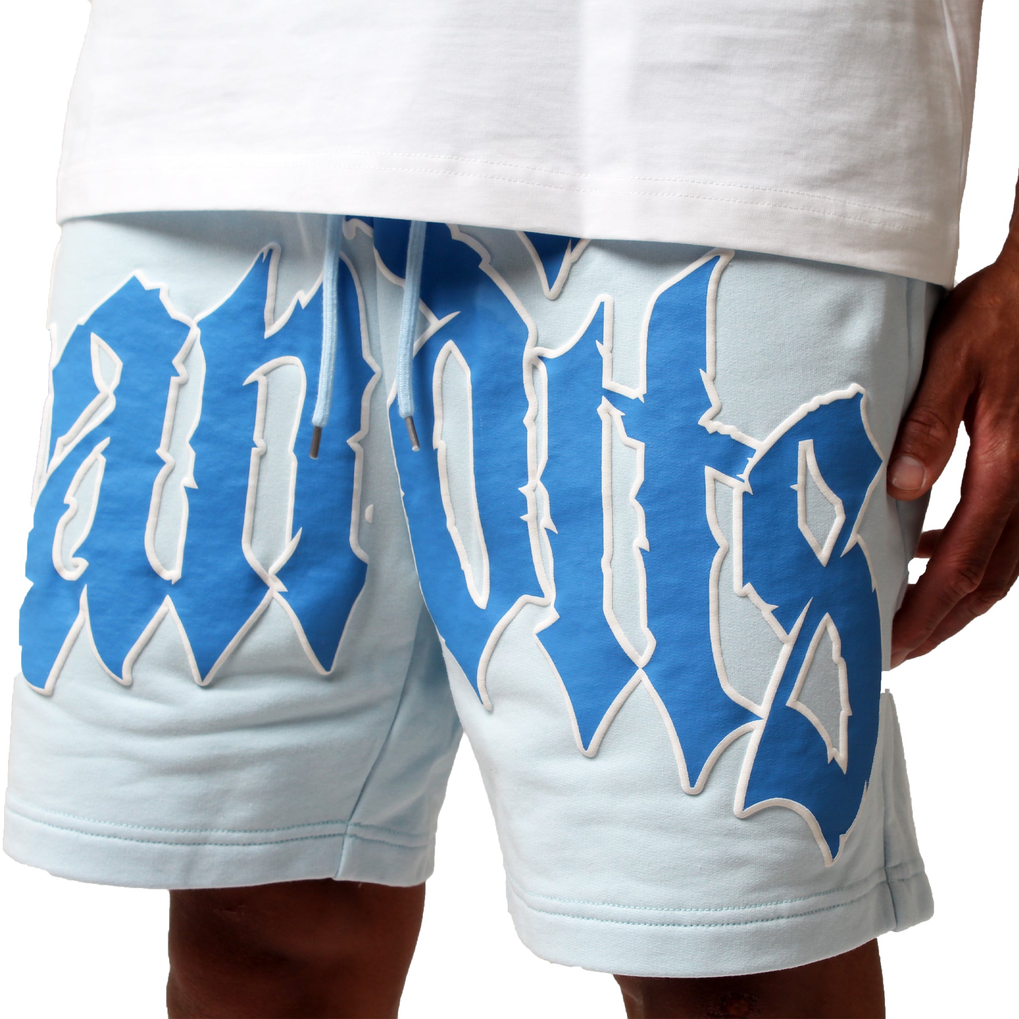 Someone is wearing the EGO SWEATSHORT LT. BLUE, featuring large, bold, blue gothic "bandits" 3D puff print across the front. The true-to-size shorts are styled with a white shirt.