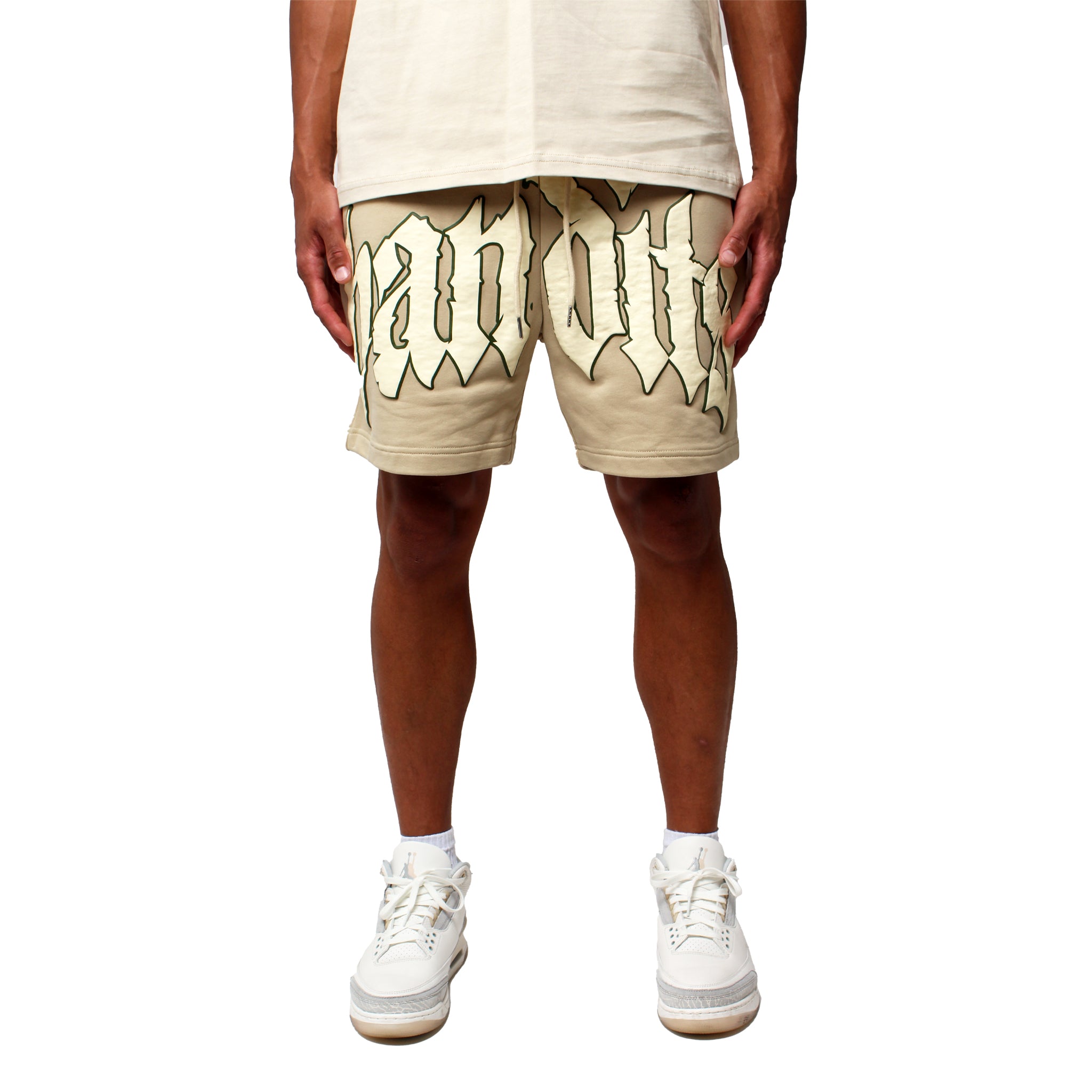A person stands against a white background wearing EGO SWEATSHORT TAN with bold black 3D Puff Print Gothic lettering, beige T-shirt, and white sneakers. The image is cropped at the torso to highlight the shorts and lower body.