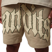 Someone wearing the EGO SWEATSHORT TAN, heavyweight French terry fleece shorts in beige, with large cream and dark green gothic 3D puff print "Bandits" text, stands with both hands on their hips.