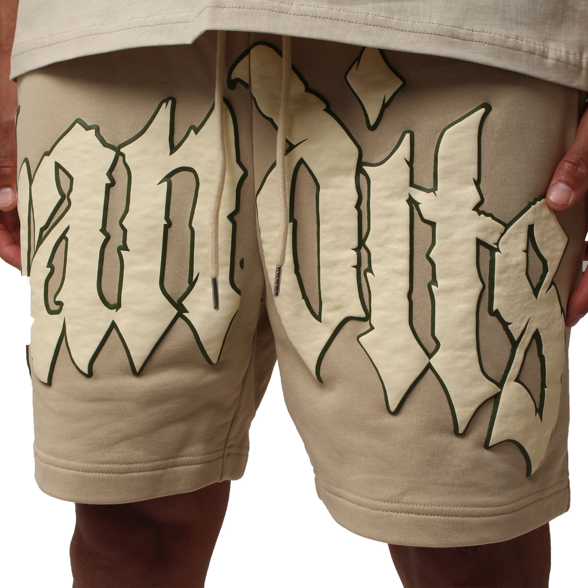 Someone wearing the EGO SWEATSHORT TAN, heavyweight French terry fleece shorts in beige, with large cream and dark green gothic 3D puff print "Bandits" text, stands with both hands on their hips.