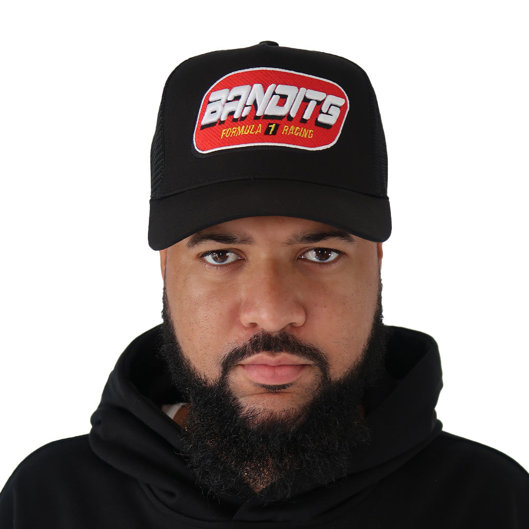 A bearded man wears a black FORMULA ONE TRUCKER hat with bold red and white 3D embroidery, paired with a black hoodie. He faces the camera against a plain white background. The adjustable snapback offers a customizable fit.