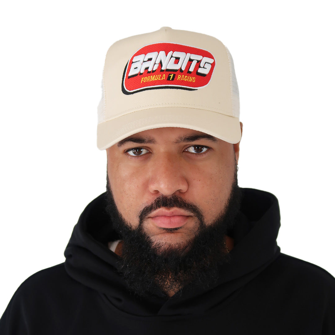 A man with a full beard and mustache wears the cream-colored FORMULA ONE TRUCKER hat with 3D embroidery and adjustable snapback, paired with a black hoodie, looking at the camera against a white background.
