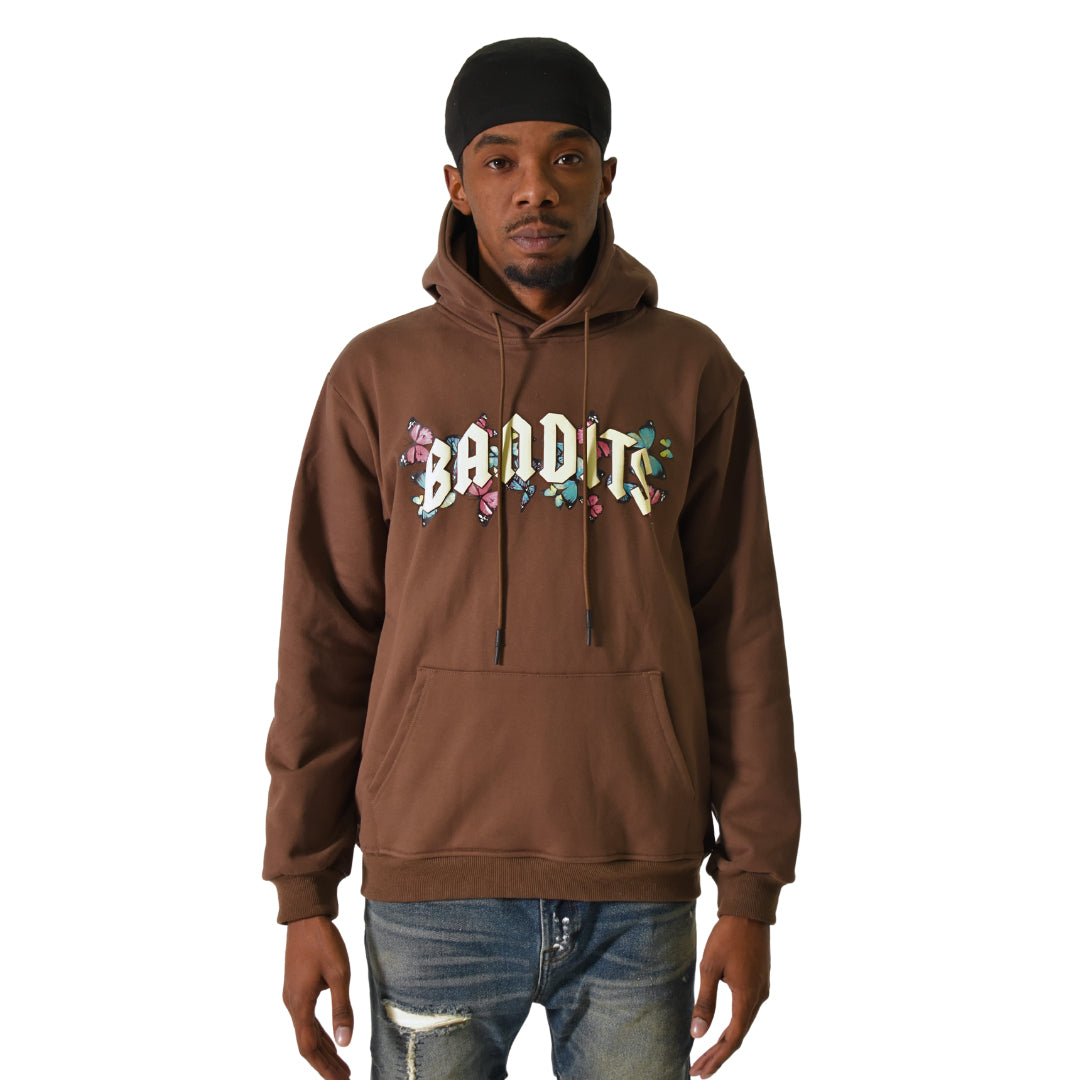 A man in a black headscarf, FLY FREE PULLOVER HOODY COCOA with BANDITS in colorful gothic lettering, and distressed blue jeans stands facing forward against a white background, highlighting bold streetwear style.