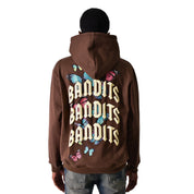 Someone shown from behind in a streetwear look, wearing the FLY FREE PULLOVER HOODY COCOA featuring "BANDITS" in gothic letters and colorful butterflies on the back, paired with blue jeans and a black cap.