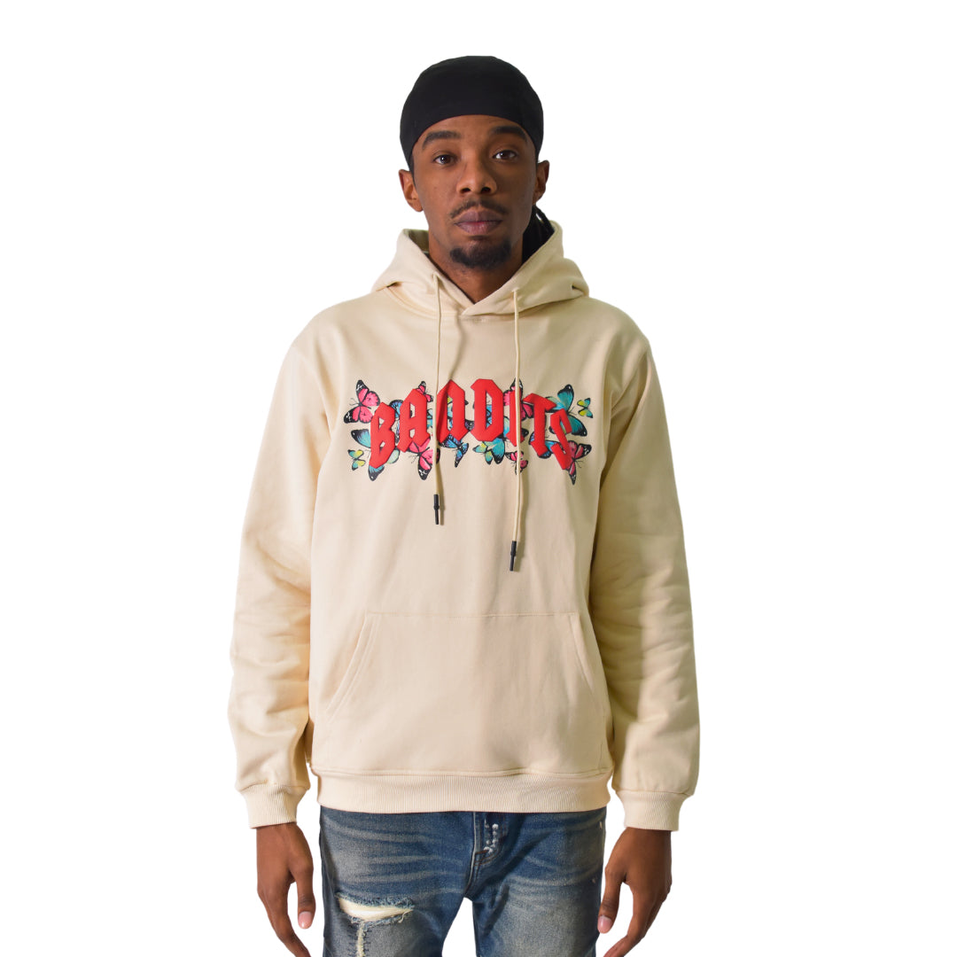 A man wears the FLY FREE PULLOVER HOODY TAN featuring a beige color, red gothic BADDIT lettering, and blue accents, paired with ripped blue jeans and a black headscarf, creating a bold streetwear look against a white background.