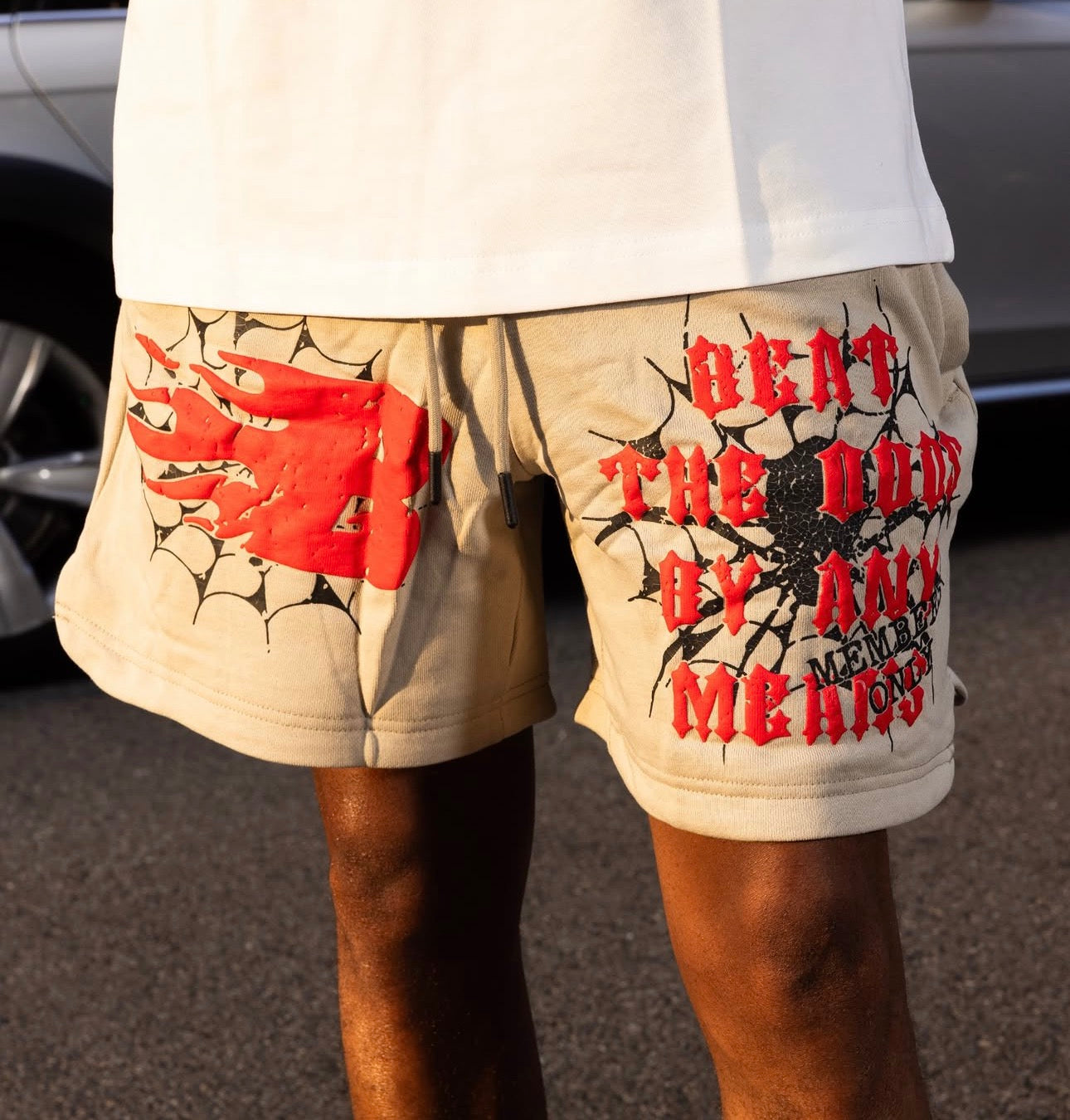 Someone wears grey BEAT THE ODDS SWEATSHORTS, crafted in heavyweight French terry fleece, featuring bold red and black graphics with a flaming emblem, web patterns, and distressed “HEAT THE DEAD OF MAY MEMORIAL” text.