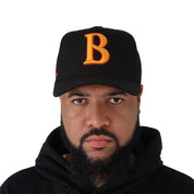 A bearded man wears a black hoodie and the GANG AINT GANG TRUCKER hat, featuring an adjustable snapback and bold orange 3D embroidered letter B, looking at the camera against a white background.