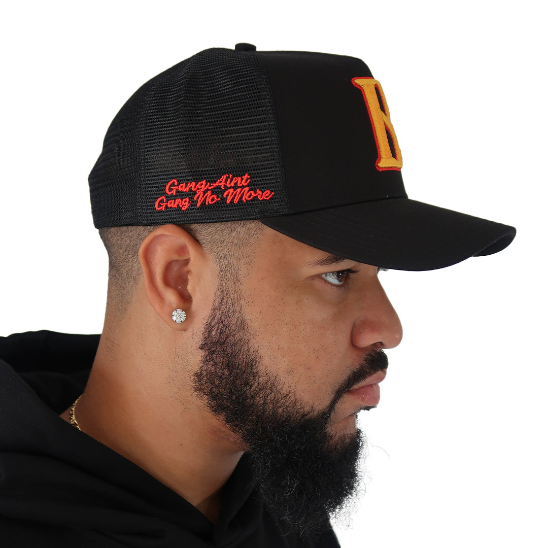 A bearded man with a diamond earring wears the GANG AINT GANG TRUCKER hat—a black mesh snapback featuring a large 3D yellow B on the front and red "Gang-Aint Gang No More" script on the side, worn turned right.