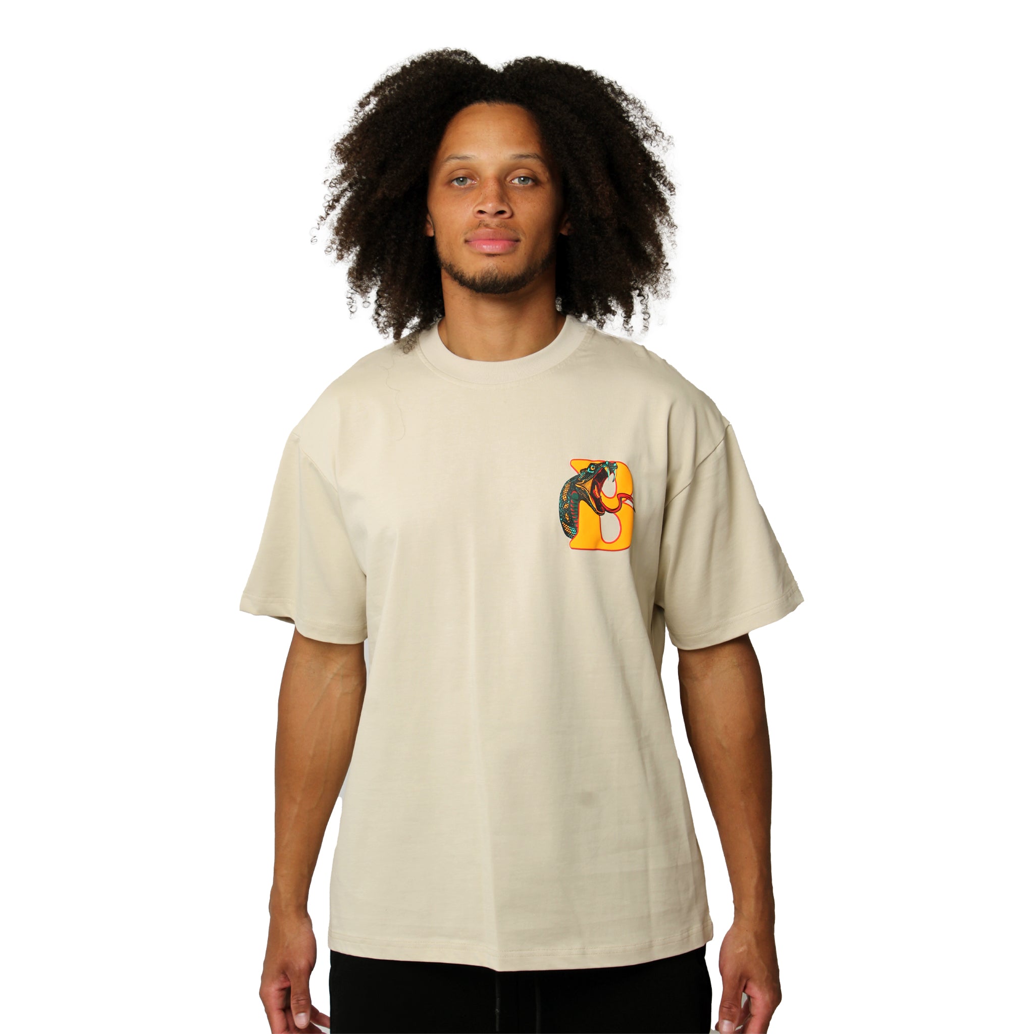 A person with long curly hair wears the GANG AIN'T GANG NO MORE S/S TEE TAN—an oversized, heavy GSM beige tee with a small colorful abstract puff print on the left chest—standing against a plain white background.