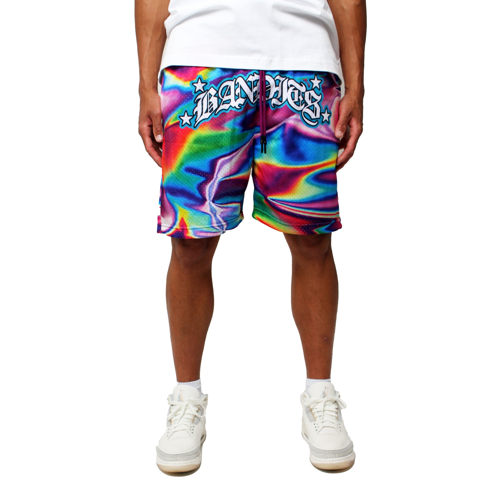 A person wearing white sneakers and a T-shirt is shown from the waist down, modeling the IRIDESCENT MESH SHORTS MLTI—colorful neon tie-dye mesh shorts with bold, stylized text and stars on the front waistband. Fits true to size.
