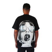 A man in a black cap and the LOLLIPOP S/S TEE BLACK stands facing away. The oversized tee features a large retro graphic of a bob-haired woman with sunglasses and a lollipop on a plain white background.