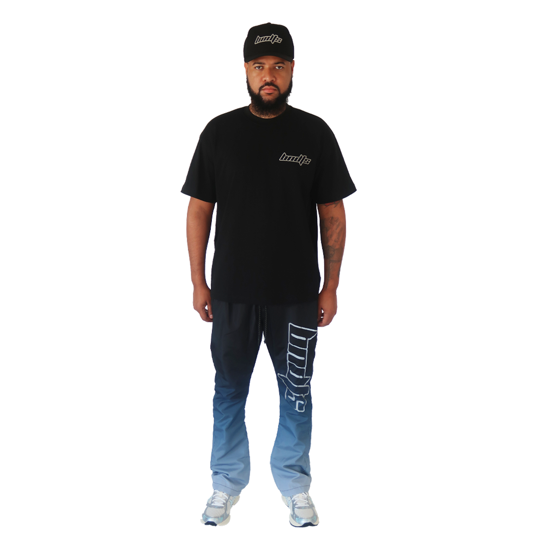 A man stands facing forward on a white background wearing the LOLLIPOP S/S TEE BLACK, a black Bandits hat, blue gradient pants with white designs, and white sneakers.
