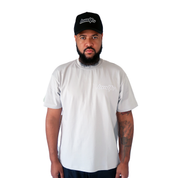 A bearded man stands facing forward, wearing a black cap and the LOLLIPOP S/S TEE GREY with a white embroidered logo. He has a tattoo on his left forearm and poses against a plain white background.
