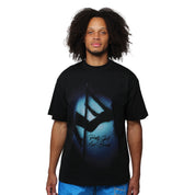 A man with curly hair faces the camera against a white background, wearing the MAIN STAGE S/S TEE BLACK—a graphic black tee with blue and black abstract design and script text—paired with blue shorts.