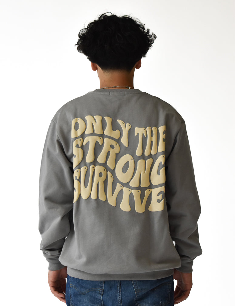 A person with curly hair, seen from behind, wears blue jeans and the ONLY THE STRONG SURVIVE CREWNECK SWEATSHIRT STONE, a gray heavyweight crewneck featuring large beige wavy text. The background is white.