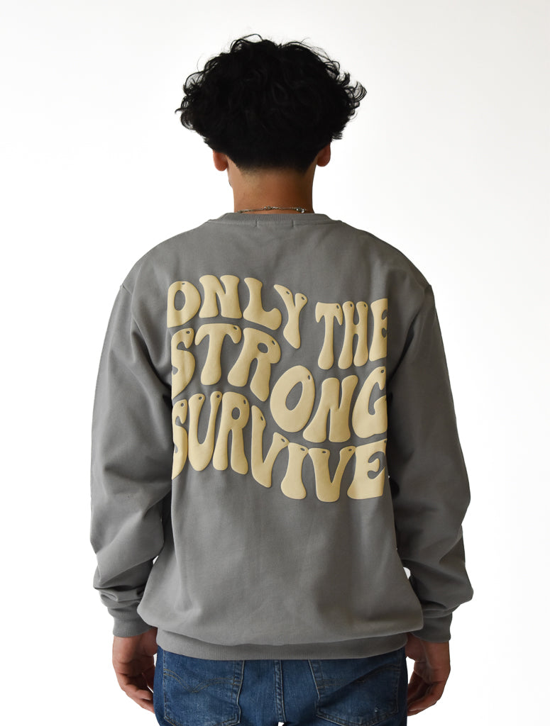 A person with curly hair, seen from behind, wears blue jeans and the ONLY THE STRONG SURVIVE CREWNECK SWEATSHIRT STONE, a gray heavyweight crewneck featuring large beige wavy text. The background is white.