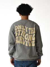 A person with curly hair, seen from behind, wears blue jeans and the ONLY THE STRONG SURVIVE CREWNECK SWEATSHIRT STONE, a gray heavyweight crewneck featuring large beige wavy text. The background is white.