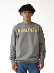 A person with curly dark hair wears the ONLY THE STRONG SURVIVE CREWNECK SWEATSHIRT STONE, a premium fleece grey heavyweight crewneck, with large cream "BANDITS" lettering, paired with blue jeans against a plain white background.