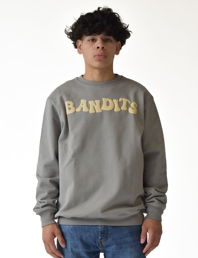 A person with curly dark hair wears the ONLY THE STRONG SURVIVE CREWNECK SWEATSHIRT STONE, a premium fleece grey heavyweight crewneck, with large cream "BANDITS" lettering, paired with blue jeans against a plain white background.