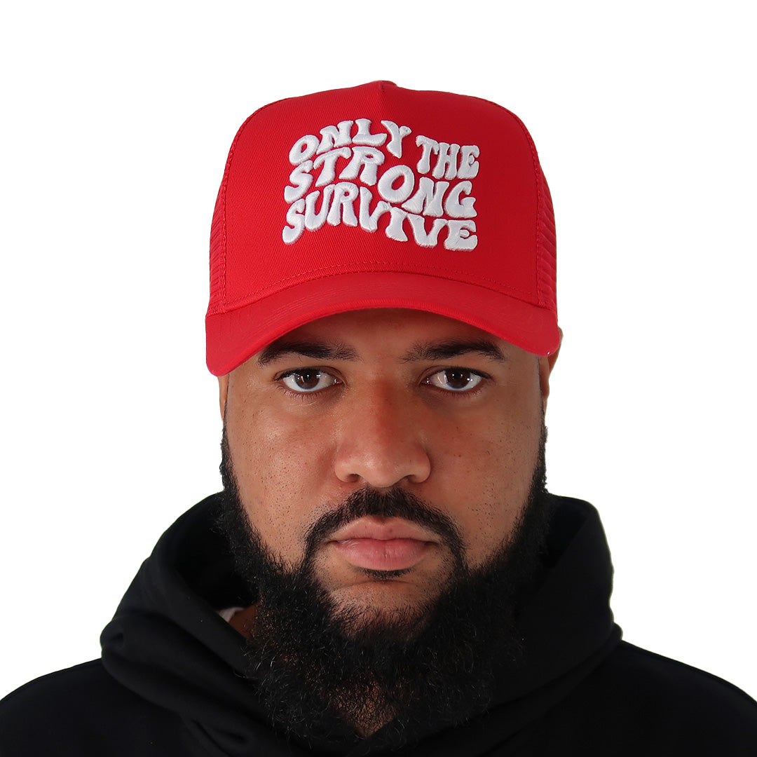 A bearded man in a black hoodie wears the ONLY THE STRONG SURVIVE TRUCKER hat, featuring bold 3D white wavy embroidery, looking directly at the camera against a white background.