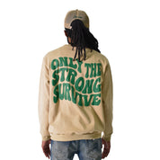 A person in a beige cap, blue jeans, and the ONLY THE STRONG SURVIVE CREWNECK SWEATSHIRT TAN featuring green 3D Puff Print text on the back, stands against a white background.