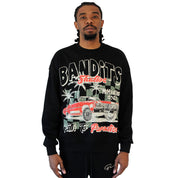 A man with braided hair wears the PATH TO PARADISE CREWNECK SWEATSHIRT BLACK, featuring a graphic of a red vintage lowrider, palm trees, and text: BANDITS, Fearless, Members Only, and Path to Paradise. He stands against a white background.