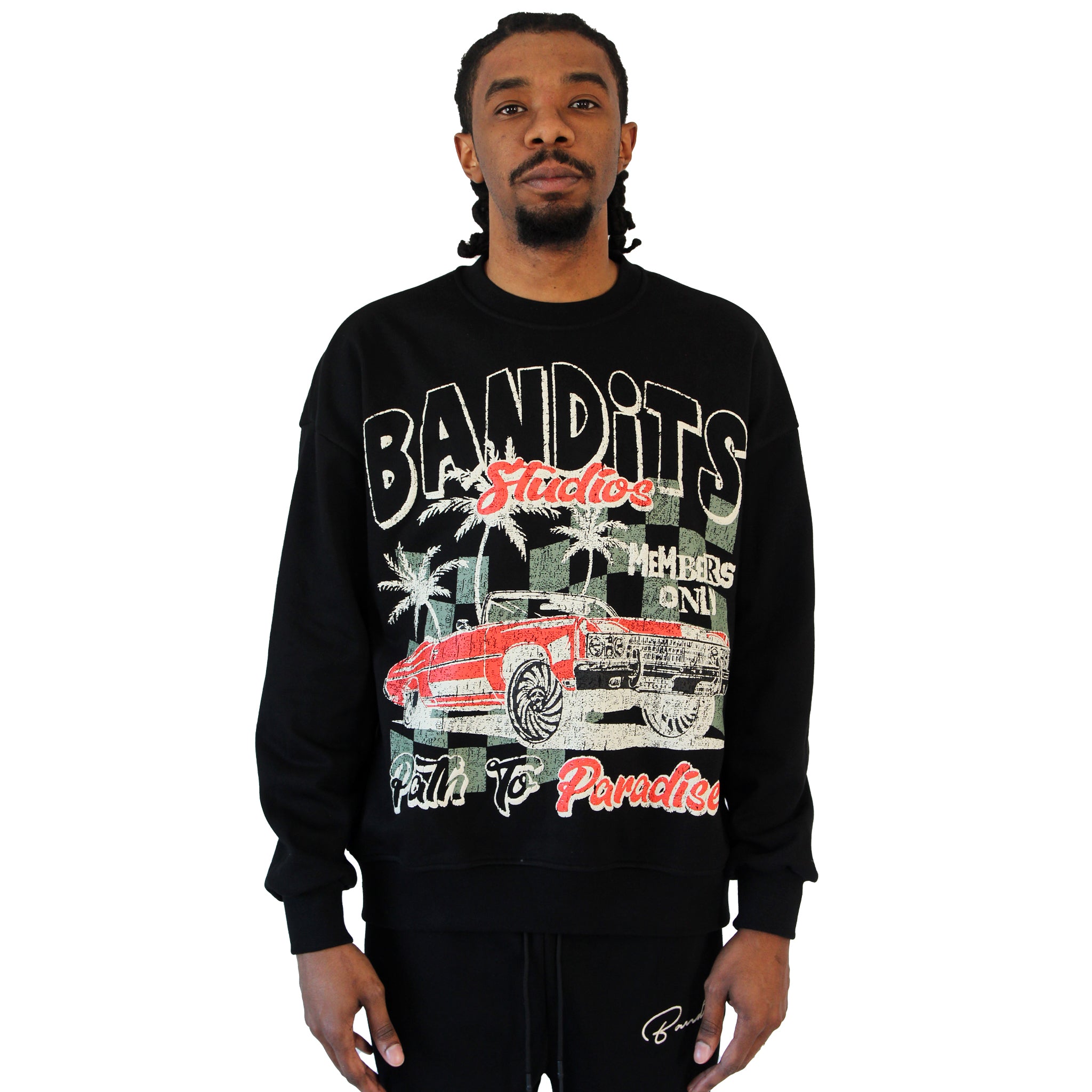 A man with braided hair wears the PATH TO PARADISE CREWNECK SWEATSHIRT BLACK, featuring a graphic of a red vintage lowrider, palm trees, and text: BANDITS, Fearless, Members Only, and Path to Paradise. He stands against a white background.