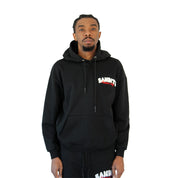 A man stands against a white background wearing the BIKER CLUB PULLOVER HOODY BLACK and matching black pants, both featuring bold white and red BANDIT lettering on the chest and thigh.