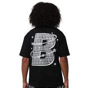 A person with curly hair faces away, wearing the VVS S/S TEE BLACK— a streetwear T-shirt with a large, patterned letter B on the back, accented by stars and orbit-like rings for a cosmic graphic tee vibe.