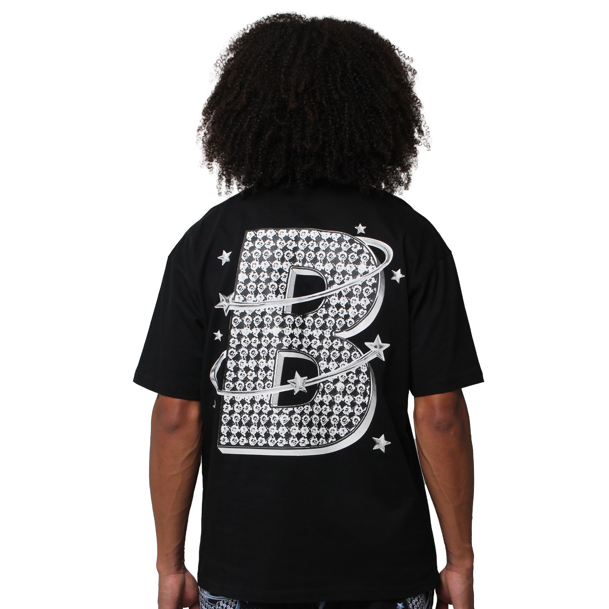 A person with curly hair faces away, wearing the VVS S/S TEE BLACK— a streetwear T-shirt with a large, patterned letter B on the back, accented by stars and orbit-like rings for a cosmic graphic tee vibe.