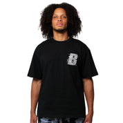 A person with curly hair wears the VVS S/S TEE BLACK, a loose black streetwear T-shirt with a white stylized "B" graphic on the left chest. The background is plain white.