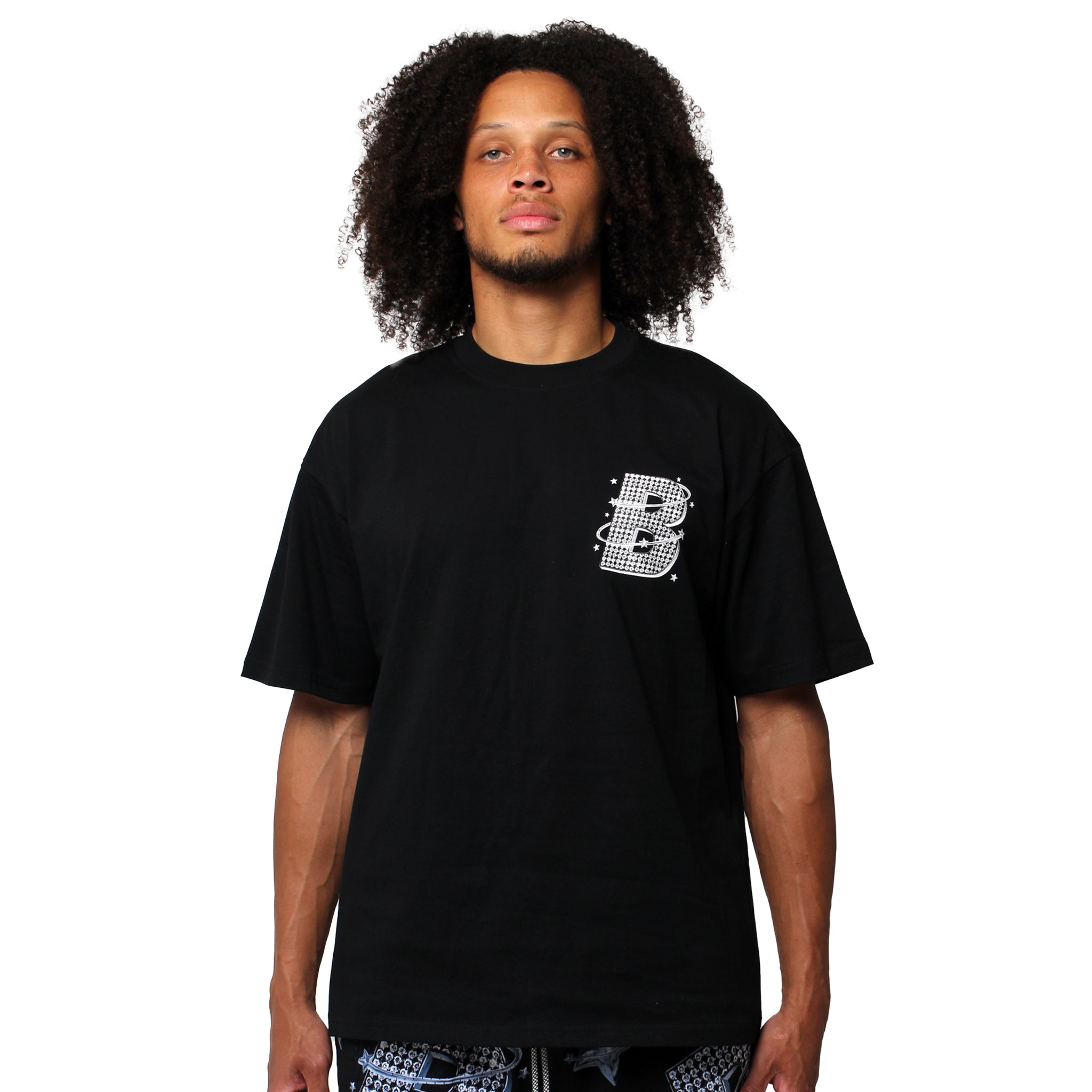 A person with curly hair wears the VVS S/S TEE BLACK, a loose black streetwear T-shirt with a white stylized "B" graphic on the left chest. The background is plain white.