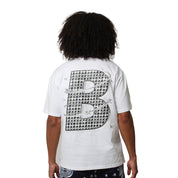 A person with curly hair is seen from behind, wearing the VVS S/S TEE WHITE, which features a large, patterned letter B and star designs on the back. The streetwear look is paired with dark shorts.