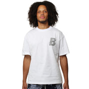 A person with curly hair wears the VVS S/S TEE WHITE, which features a small, patterned letter B on the upper left side, standing against a plain white background.
