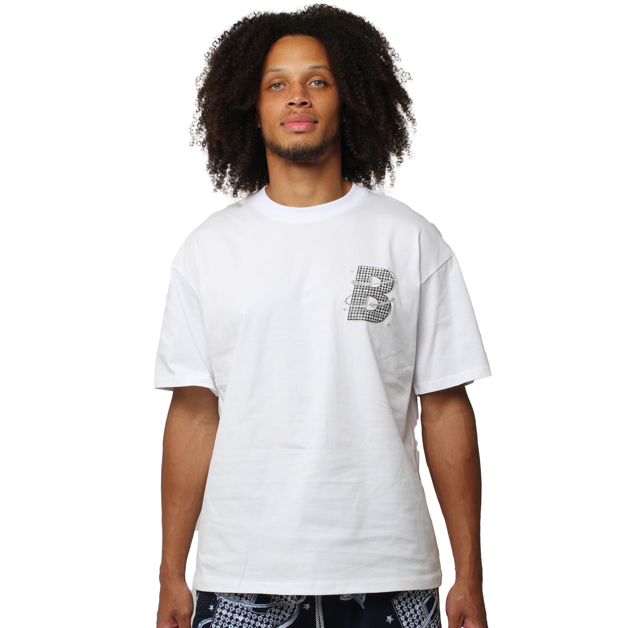 A person with curly hair wears the VVS S/S TEE WHITE, which features a small, patterned letter B on the upper left side, standing against a plain white background.