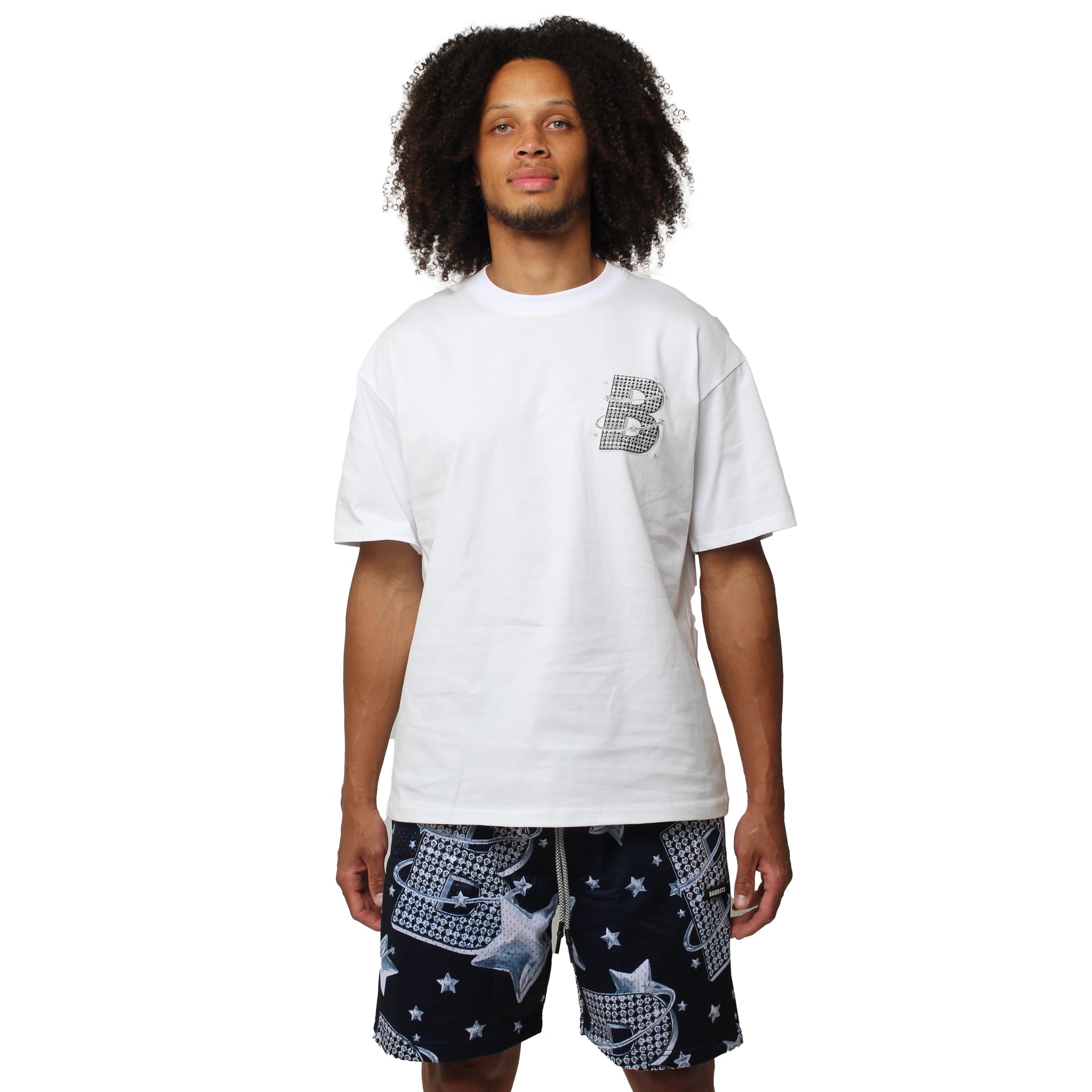 A man with curly hair wears the VVS S/S TEE WHITE, a loose streetwear T-shirt with a small checkered B logo on the chest, and black shorts with star and net patterns, standing against a plain white background.