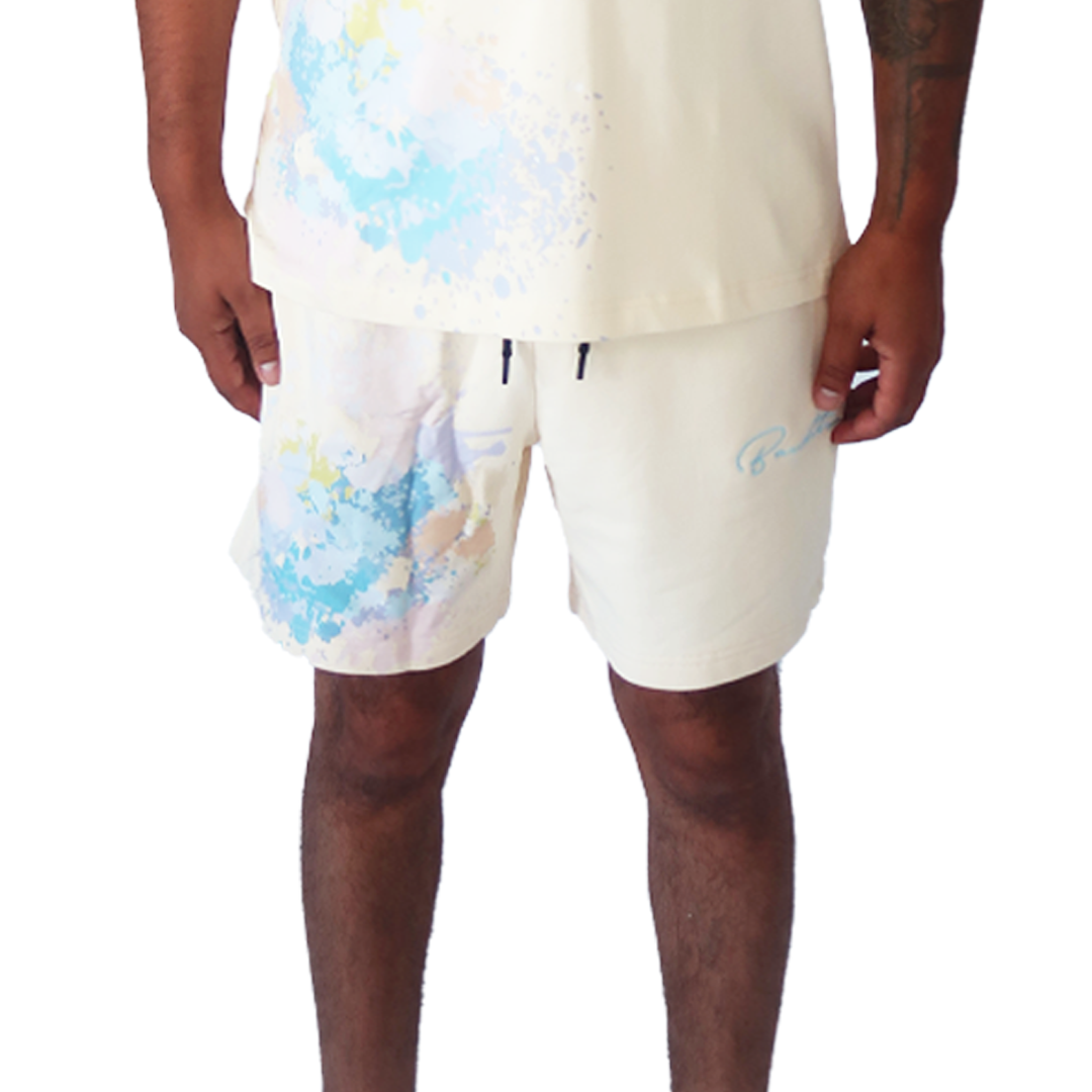 Someone stands with arms relaxed by their sides against a plain white background, wearing the WORK OF ART SWEATSHORTS CREME—oversized, light-colored shorts featuring pastel watercolor splashes and a matching shirt.