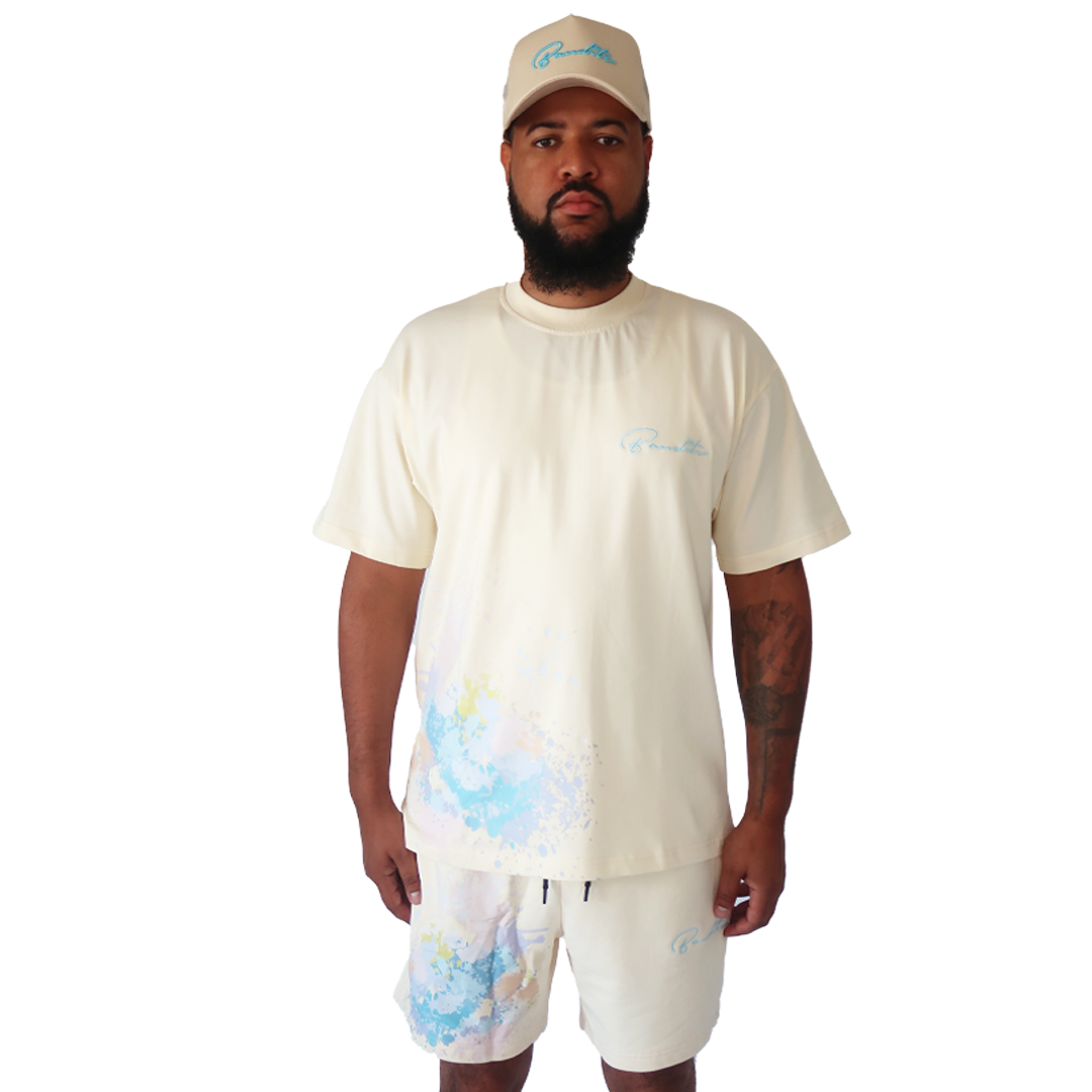 A man stands facing forward against a white background, wearing the WORK OF ART S/S TEE CREME—an oversized beige heavy GSM cotton tee with subtle embroidery—and matching shorts and cap featuring blue and yellow splatter designs.