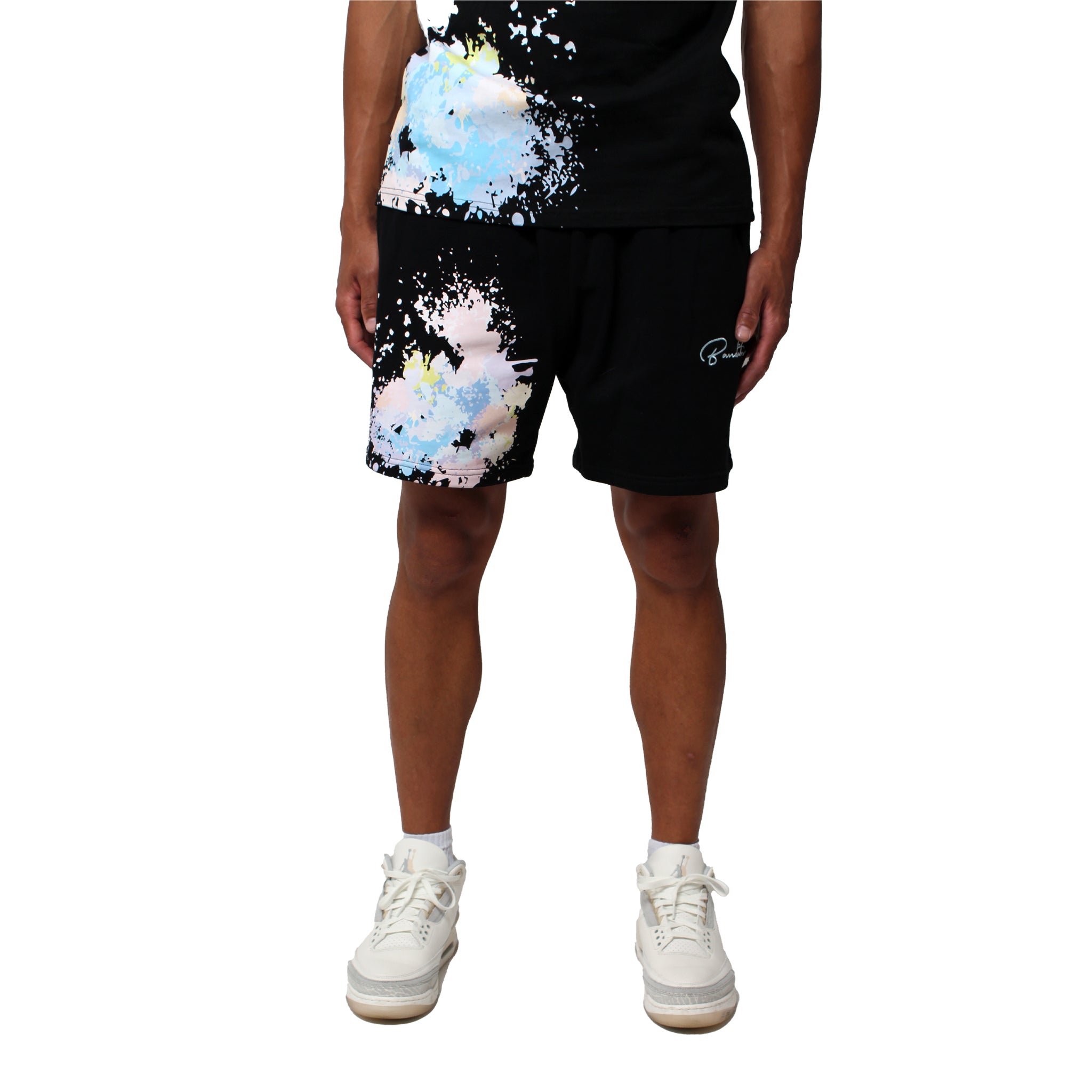 A person wears WORK OF ART SWEATSHORTS BLACK—oversized, heavyweight black sweatshorts—and a black tee with blue, white, and pink paint splatter embroidery. The background is white and their face is not visible.