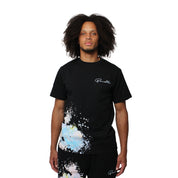 Wearing the WORK OF ART S/S TEE BLACK, a person with curly hair and matching shorts stands against a plain white background. The oversized black T-shirt features a vibrant paint splatter design.